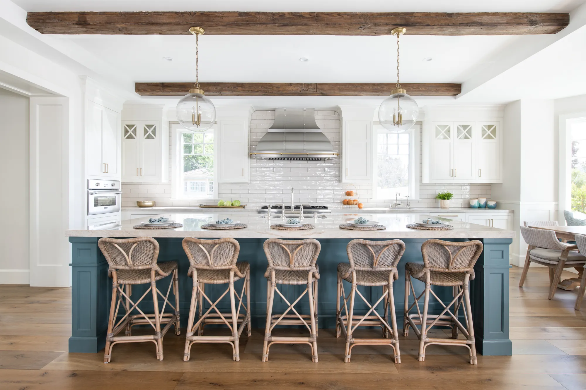 White shaker cabinetry and a stainless range hood wrapping a large blue island with woven counter stools in a wide coastal kitchen.