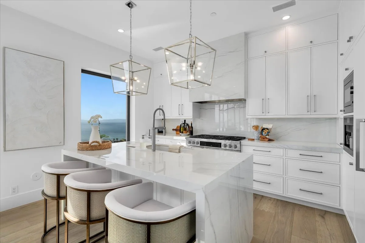 Waterfall-edge island and lantern pendants in a white marble kitchen framed by an ocean-view window, kitchen-specialist work start to finish.