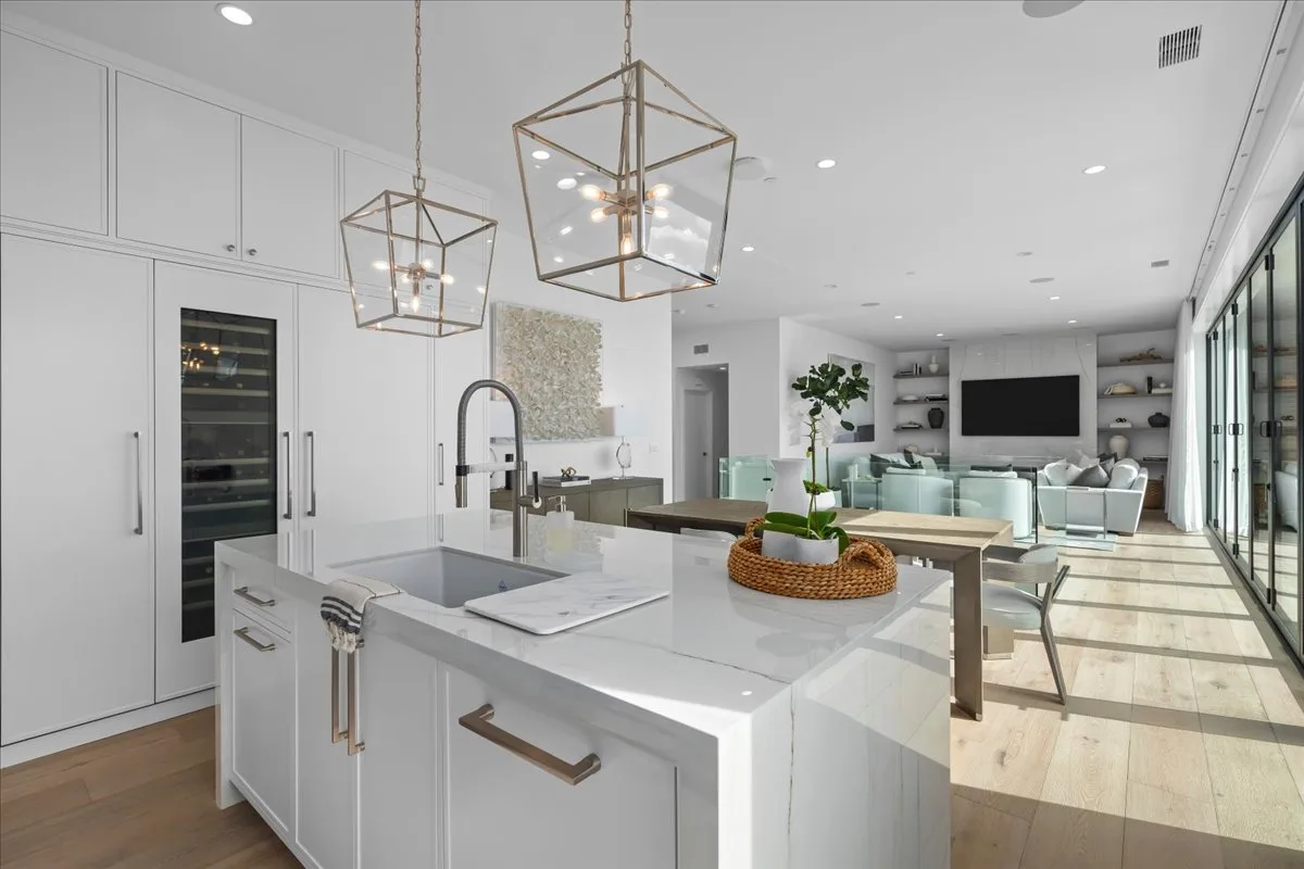 Marble island with a prep sink anchors an open-plan white kitchen that opens to an indoor-outdoor living space.