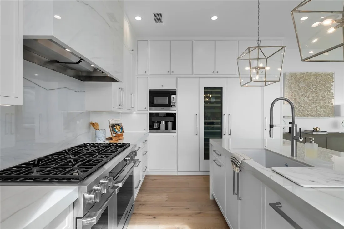 A full-height slab backsplash rises behind a professional stainless range and custom hood beside a waterfall island and built-in wine fridge.